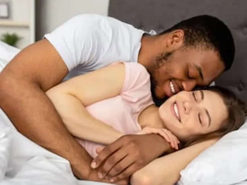 A clothed couple cuddling and smiling together on a bed in warm light
