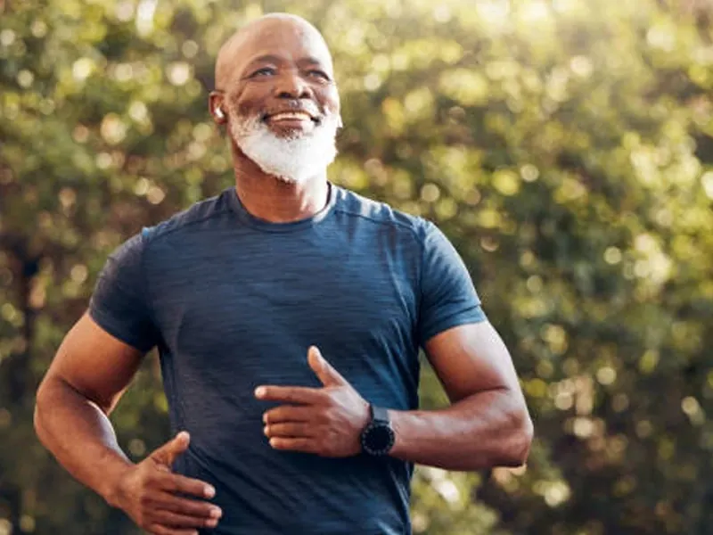 A smiling older man in a blue T-shirt jogging outdoors among trees (brand stock photo, model)